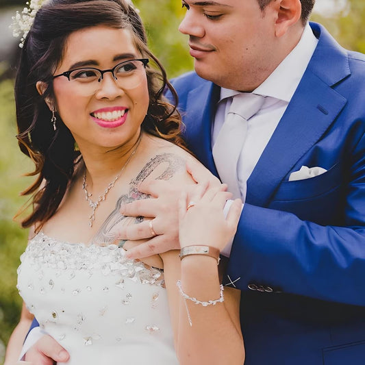 Groom in a blue suit embraces the bride, who is wearing a strapless embellished white gown and a delicate leaf-shaped necklace. She smiles brightly, showing a tattoo on her shoulder, with matching bracelet and earrings completing her look.