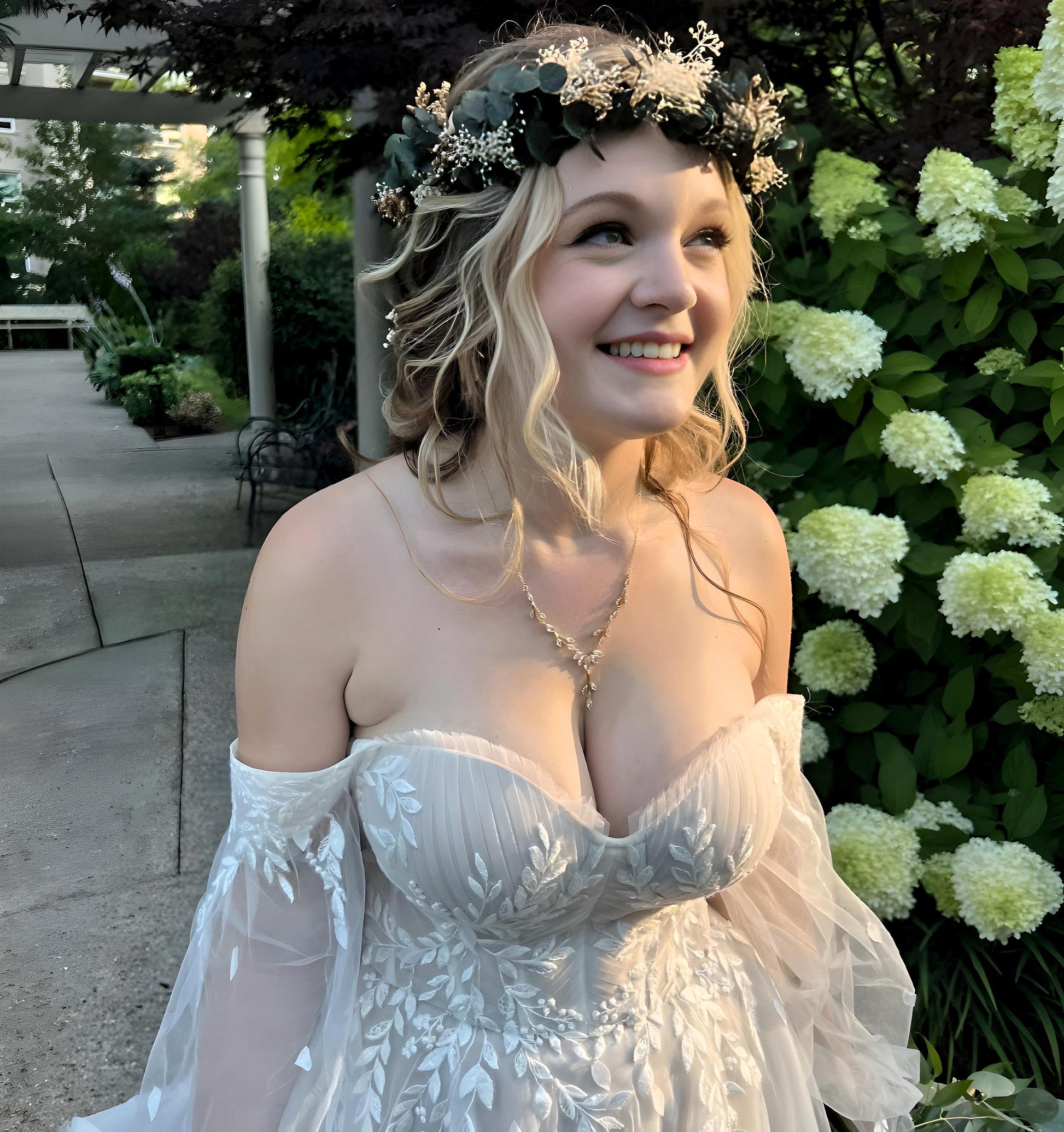 Smiling woman in a white lace off-shoulder gown wearing a delicate leaf-shaped necklace, standing outdoors with a flower crown. White hydrangeas and greenery are in the background.