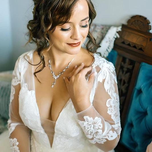 Bride wearing a long-sleeved lace wedding dress, showcasing a sparkling leaf-shaped necklace with crystal details. She is seated on a vintage chair, gently touching the necklace with a serene expression.