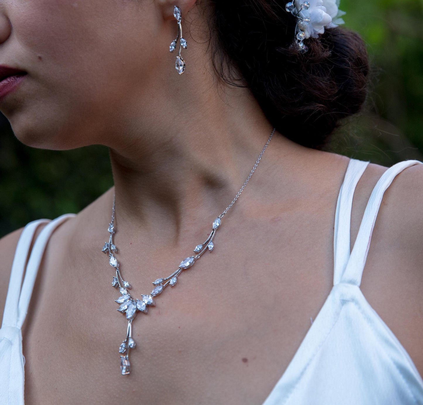 Close-up of a woman wearing a silver leaf-shaped crystal necklace and matching dangling earrings, paired with a white satin dress. The jewelry features delicate floral details, perfect for bridal or formal occasions.